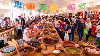 Mujeres con delantales sirven mole desde cazuelas de barro en una carpa adornada con tiras de papel picado y puestos de comida.