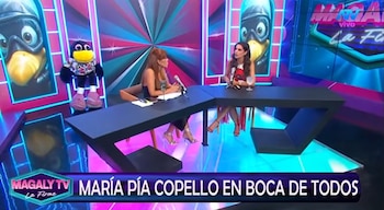 Dos mujeres sentadas en una mesa angular con micrófonos, vasos, y un muñeco de pájaro. Fondo con figura de ave animada y luces de neón