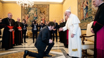 El presidente Javier Milei, vestido de traje oscuro, arrodillado ante el Papa Francisco, quien viste de blanco, en un salón del Vaticano con tapices y otras personas.