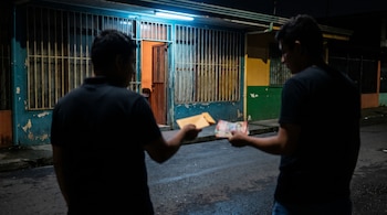 Dos figuras de espaldas y desenfocadas intercambian un sobre y un fajo de billetes en una calle oscura frente a un local con rejas y puerta entreabierta.