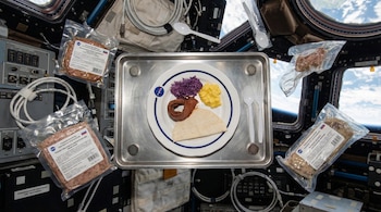 Un plato blanco con logo NASA, repollo, maíz, tortilla y carne en bandeja magnética en la ISS, con envases de comida al vacío y utensilios flotando.