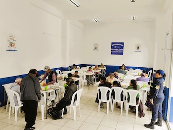 Vista interior de un comedor con mesas y sillas blancas. Numerosas personas sentadas comiendo. Hay un cartel de "Bienvenidos Comedor Social" en la pared