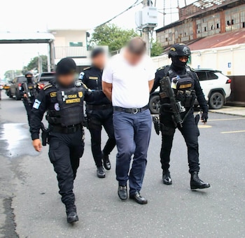 Agentes policiales con chalecos tácticos escoltan a un hombre esposado con camiseta blanca y pantalón oscuro en una calle
