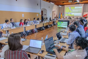 Una sala de conferencias con mesas largas, personas sentadas, banderas de países, laptops, botellas de agua y una pantalla con imagen de naturaleza
