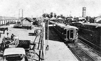 Fotografía en blanco y negro de una estación de tren con múltiples vías, varios vagones, un andén con personas, carruajes de caballos y un coche antiguo