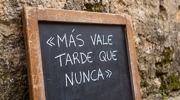 Una pizarra de madera apoyada en una pared de piedra con el texto «MÁS VALE TARDE QUE NUNCA» escrito en tiza blanca.