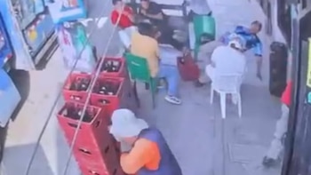 Varias personas en un espacio exterior, algunas sentadas, una caída. Hay cajas rojas apiladas con botellas y una persona agachada cerca de ellas