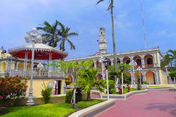 Plaza con un kiosco de techo rojo, un edificio con arcos y un campanario con reloj, varias palmeras, farolas blancas y jardines verdes