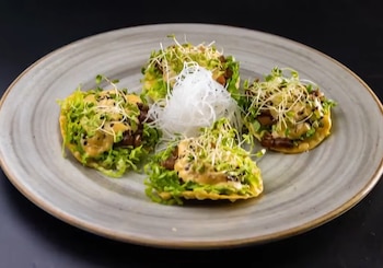 Cuatro porciones de comida sobre un plato gris redondo, decoradas con lechuga, salsa y brotes verdes, dispuestas alrededor de un adorno blanco