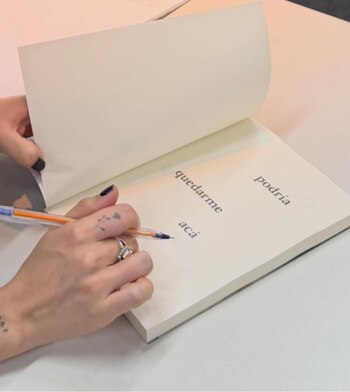 Primer plano de manos femeninas con anillo y uñas oscuras escribiendo en un libro abierto con un bolígrafo. Las palabras 'quedarme' y 'acá' son visibles