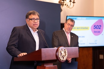 Un hombre de lentes y traje oscuro pronuncia un discurso en un podio con emblema, con su mano derecha sobre él. Detrás, otro hombre y una pantalla muestran '502 becas'
