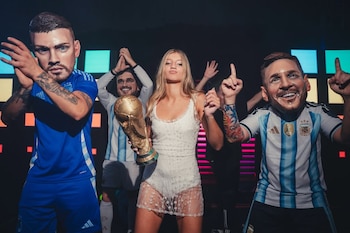 Allegra Cubero, una joven rubia vestida de blanco, sostiene una réplica de la Copa del Mundo, flanqueada por figuras con cabezas grandes de jugadores de fútbol