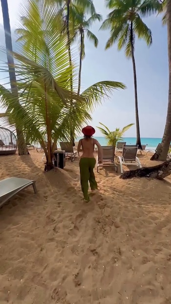 Una mujer con sombrero rojo y pantalón verde camina de espaldas por la arena entre palmeras y reposeras hacia el mar