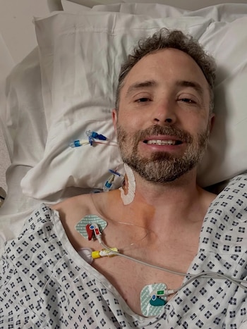Hombre barbudo con pelo rizado y bata de hospital yace en una cama blanca, con catéteres y electrodos visibles en su pecho y cuello