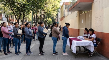 Una fila de mujeres mexicanas, algunas con niños o documentos, espera frente a una escuela de tonos crema y terracota. Dos funcionarias con chalecos guindas atienden en una mesa revisando papeles.