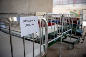 Cama de hospital metálica en primer plano con cartel "TRASLADAR A DEPOSITO". Varias camas similares con colchones blancos se extienden al fondo