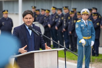 Un hombre con saco oscuro habla ante un micrófono en un atril con varios oficiales uniformados alineados al fondo