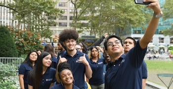 Un grupo de jóvenes con camisetas de cuello azul se toma una fotografía con un teléfono móvil en un parque urbano con edificios al fondo