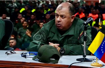 Oficial militar de uniforme verde oliva con parches FANB y REDI Los Llanos sentado en una mesa con micrófono, bandera de Venezuela, gorra y lentes