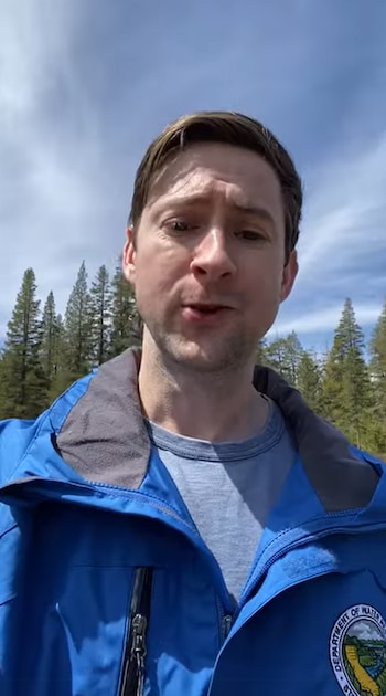 Un hombre viste una chaqueta azul con un emblema circular frente a un entorno de árboles y cielo con nubes
