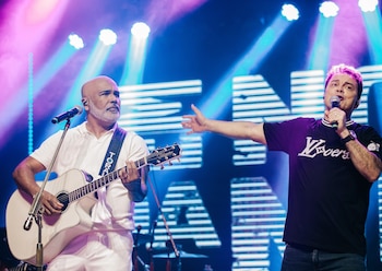 Dos hombres cantan en un escenario iluminado; uno toca la guitarra acústica y el otro sostiene un micrófono con el brazo extendido