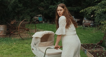 Una mujer de cabello largo y vestido blanco empuja un coche de bebé beige en un exterior. Al fondo hay árboles, chatarra y vehículos viejos