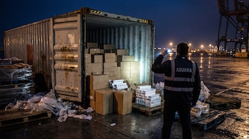 Un agente de aduanas de espaldas ilumina con una linterna el interior de un contenedor abierto con cajas de cartón en un muelle nocturno.