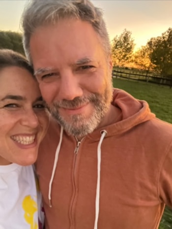 Rostros de un hombre y una mujer sonriendo, con el hombre en primer plano. Él viste una sudadera naranja con cierre, barba y cabello canoso. El fondo muestra un paisaje rural al atardecer