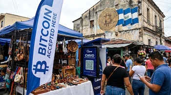 Vista de un mercado callejero en El Salvador con un kiosco de Chivo Wallet, un letrero que dice 'BITCOIN ACCEPTED HERE' y un mural de Bitcoin y la bandera salvadoreña.