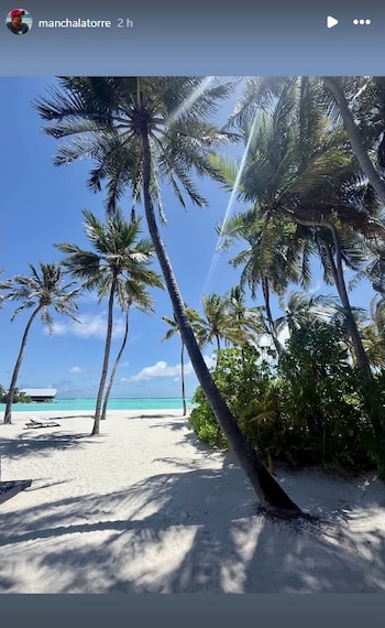 Hermosa playa de arena blanca con palmeras. Mar azul turquesa bajo un cielo despejado. El sol brilla, proyectando sombras. Un bungalow sobre el agua se ve