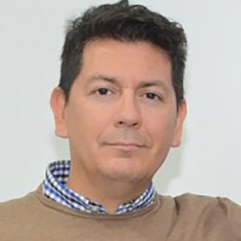Primer plano de un hombre de mediana edad con cabello oscuro, suéter beige y camisa a cuadros azul y blanco. Fondo blanco