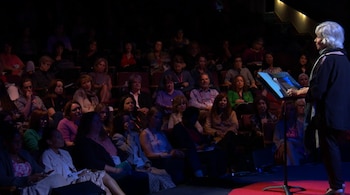 Una mujer de cabello canoso habla desde un atril en un escenario frente a un público sentado en un auditorio a oscuras