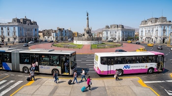 Vista aérea de la Plaza Dos de Mayo con autobuses del Metropolitano y Corredor Rosado en el primer plano, pasajeros en aceras y un monumento central