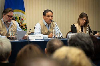 Tres miembros de la MOE/OEA, incluyendo a Gerardo de Icaza y Víctor Rico, sentados en una mesa con micrófonos y vasos, con la bandera de la OEA de fondo, presentan un informe a una audiencia