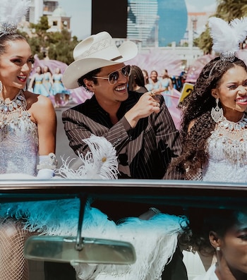 Bruno Mars en un sombrero vaquero y gafas de sol, sonriendo y señalando, en un convertible azul con dos showgirls vestidas de blanco brillante con plumas