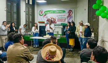 Hombre con camisa blanca habla al micrófono frente a un público que aplaude. Detrás, un banner de campaña y globos verdes decoran el evento