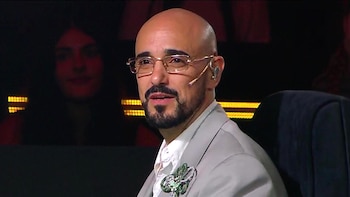 Primer plano de Abel Pintos, un hombre calvo con barba de chivo y gafas doradas, mirando hacia la derecha. Viste traje claro con broche verde y un auricular. Fondo oscuro con luces borrosas