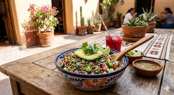 Ceviche de lentejas en tazón azul sobre mesa de madera. Incluye aguacate, cilantro, lima, salsa verde y bebida roja en un patio soleado