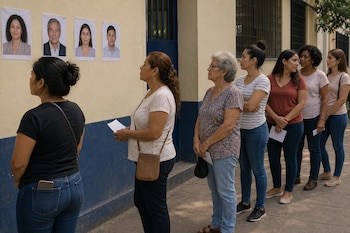 Fila de mujeres en una acera junto a una pared donde cuelgan cuatro retratos impresos de candidatos.