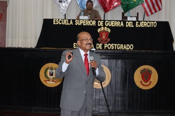 Hombre de traje con micrófono y control. Detrás, cartel "Escuela Superior de Guerra del Ejército" con texto dorado, dos escudos y banderas