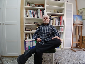 Hombre mayor de cabello y barba blancos, con camisa a cuadros azul y verde, sentado en un taburete. Detrás hay una estantería con libros y carpetas