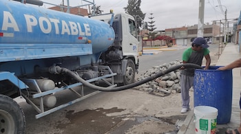 Un camión cisterna azul surte agua a un hombre que sostiene una manguera conectada a un barril de plástico en la calle.