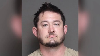 Mugshot de un hombre caucásico con pelo oscuro y barba. Tiene ojos verdes y viste una camiseta clara. Mira fijamente a la cámara con expresión neutra
