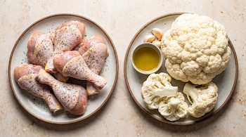 Dos platos cerámicos con muslos de pollo crudos condimentados, una coliflor, trozos cortados, dientes de ajo y un cuenco con aceite de oliva.