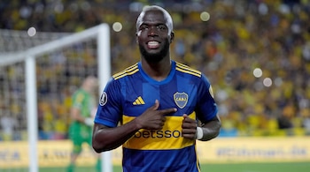 Futbolista de piel oscura y cabello claro, con camiseta azul y amarilla de Boca Juniors, señala el escudo del club en un estadio de fútbol.