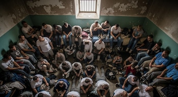 Un grupo de hombres y una mujer en una celda con paredes descascaradas, una ventana enrejada, sentados en el suelo y en camastros estrechos.