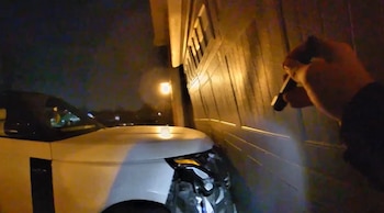 Vista nocturna de un coche blanco dañado, con el faro roto, estacionado contra una puerta de garaje de madera. Una mano sostiene una linterna iluminando la escena