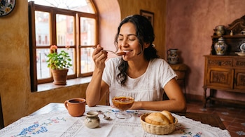 Una mujer frente a una mesa con un recipiente con cajeta, una taza de barro, pan en una cesta y una ventana colonial.