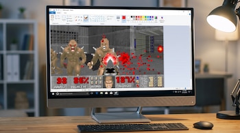 Un computador de escritorio muestra la interfaz de Microsoft Paint con un fotograma del videojuego Doom. Hay un teclado, un mouse y una lámpara en la mesa.