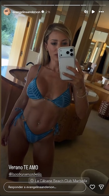 Mujer rubia de pie tomándose una selfie en un espejo, viste bikini azul con estampado y detalles dorados. Fondo con muebles de madera y espejos